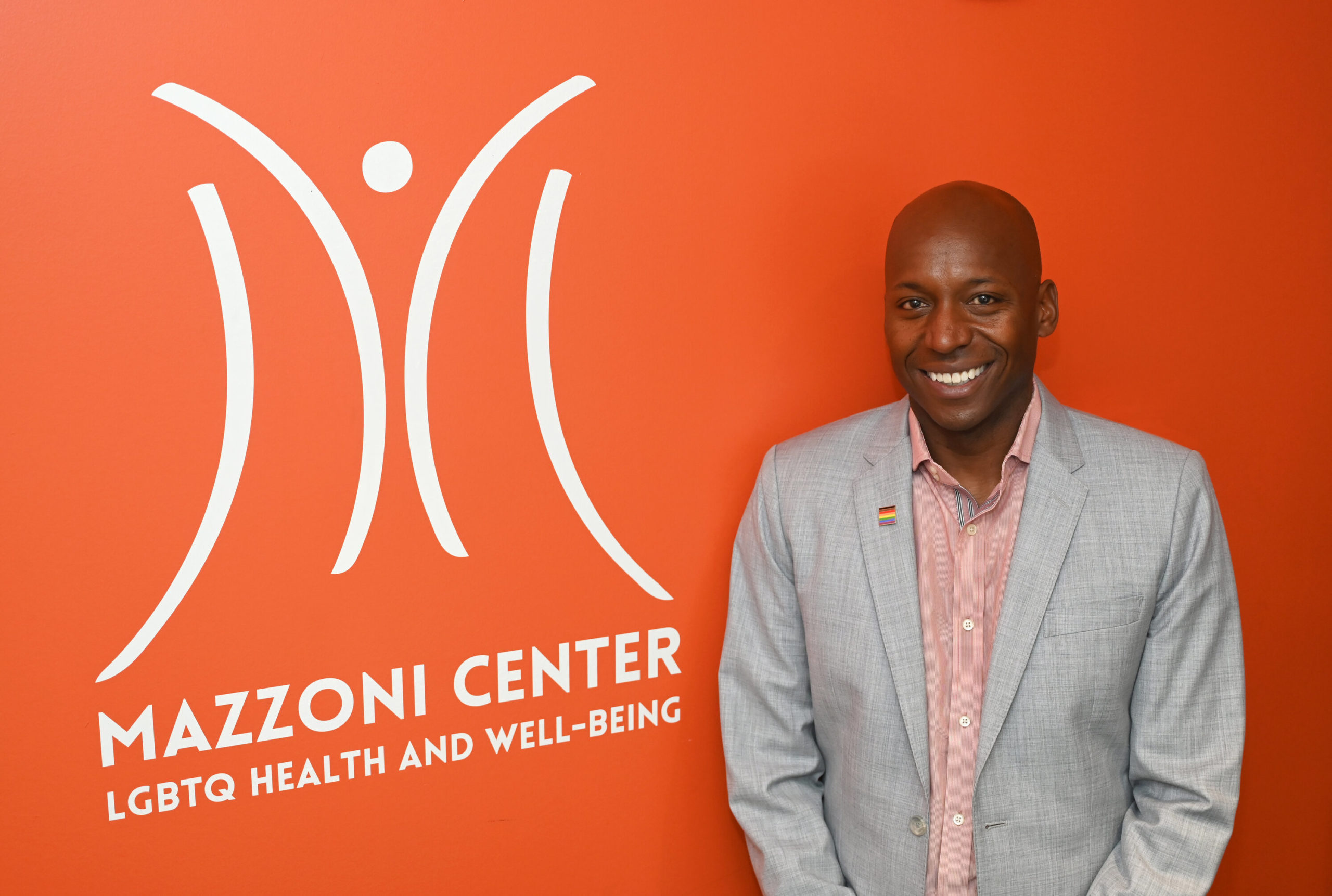 Sultan Shakir, the newest President and Executive Officer of the Mazzoni Center, standing in front of an orange wall with the Mazzoni Center logo. Underneath the logo is text that reads: Mazzoni Center LGBTQ Health and Well-Being.